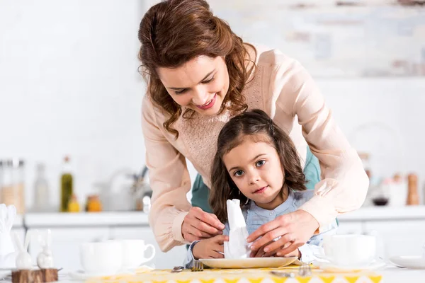 Улыбающаяся мать и маленькая дочь складывают салфетки на кухне — стоковое фото