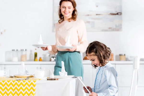 Enfant utilisant une tablette numérique pendant que la mère sert une table dans la cuisine — Photo de stock