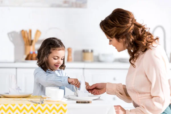 Curly mother and daughter folding napkins at table in kitchen — Stock Photo