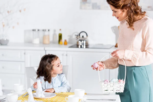 Mujer rizada sosteniendo canasta de mimbre con huevos de Pascua y mirando a su hija en la cocina - foto de stock