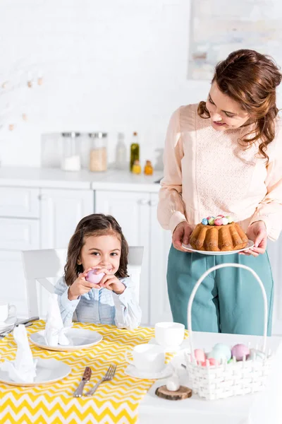 Kleines Kind hält bemaltes Ei in der Hand und blickt in die Kamera, während Mutter den Ostertisch serviert — Stockfoto