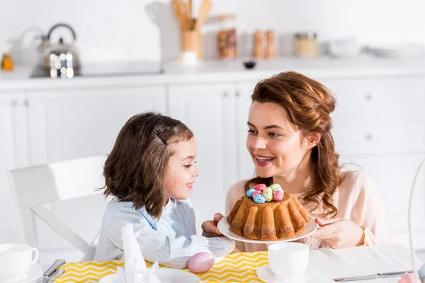 Mother showing easter cake with painted eggs to daughter in kitchen — Stock Photo