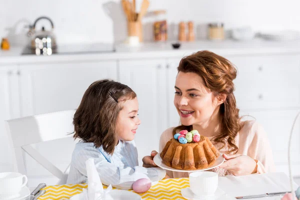 Madre mostrando pastel de Pascua con huevos pintados a su hija en la cocina - foto de stock