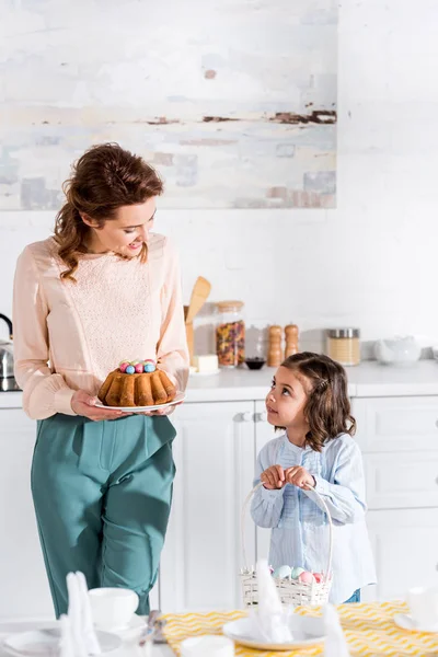 Niño con cesta de mimbre y madre con pastel de Pascua mirándose en la cocina - foto de stock