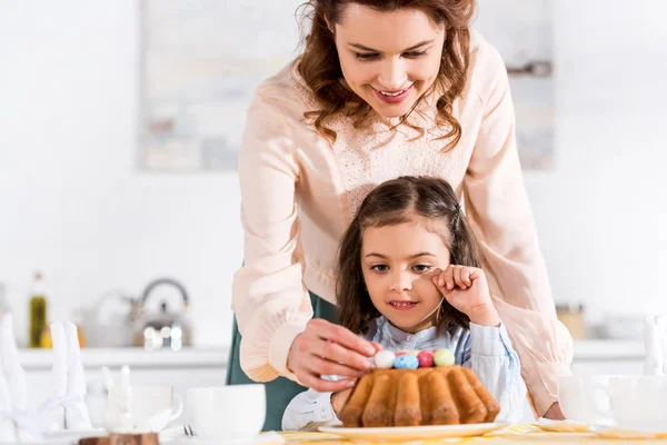 Усміхнена мати і маленька дочка прикрашають великодній торт з розфарбованими яйцями — стокове фото