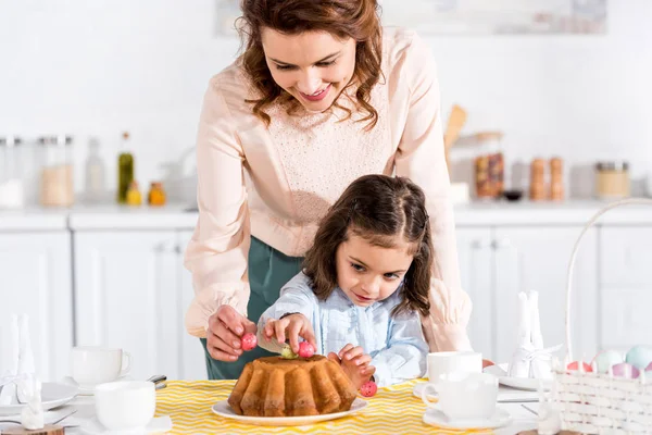 Smiling mother and daughter decorating easter cake with eggs in kitchen — Stock Photo