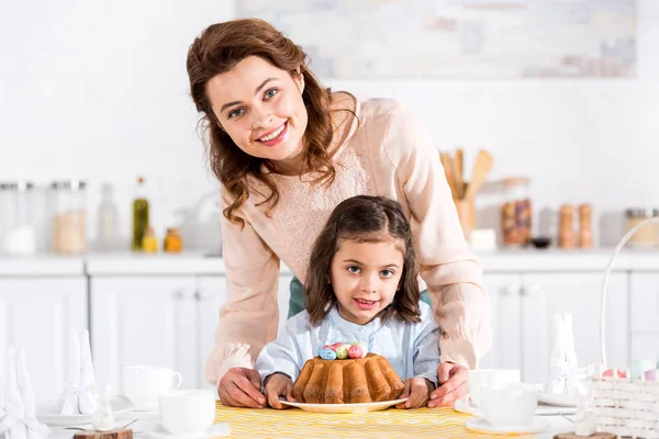 Mãe sorridente e criança com bolo de Páscoa olhando para a câmera na cozinha — Fotografia de Stock