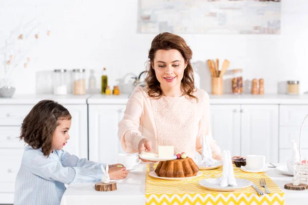 Mother and daughter sitting at table served with easter cake, napkins and ceramic rabbits — Stock Photo