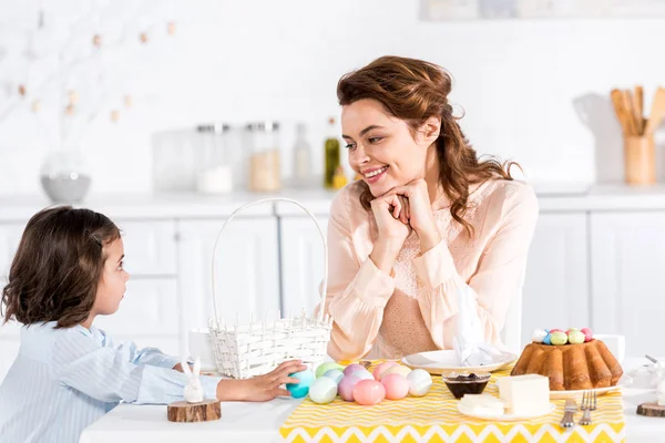 Мать и ребенок сидят за столом с пасхальным тортом и крашеными яйцами и смотрят друг на друга — стоковое фото