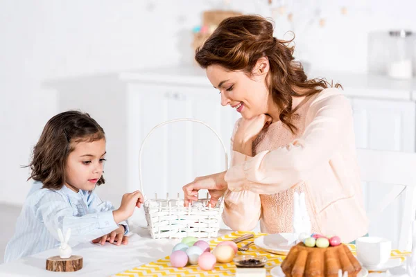 Мать и дочь кладут пасхальные яйца в плетеную корзину на кухне — стоковое фото