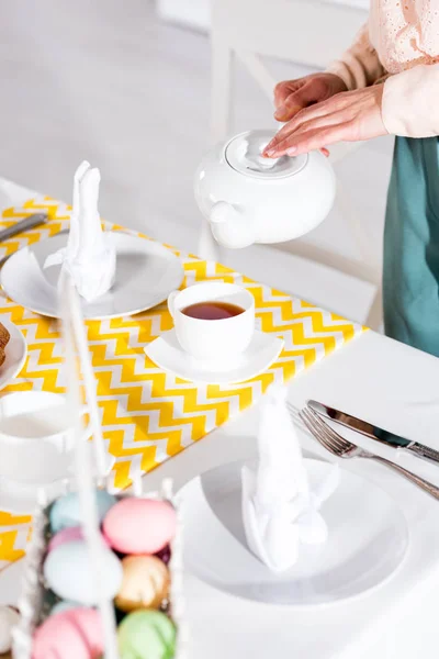 Vista parcial de la mujer con la tetera vertiendo té en la taza sobre la mesa - foto de stock