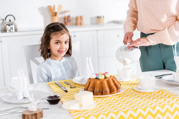 Sonriente niño mirando a la cámara mientras la madre vierte té en taza - foto de stock