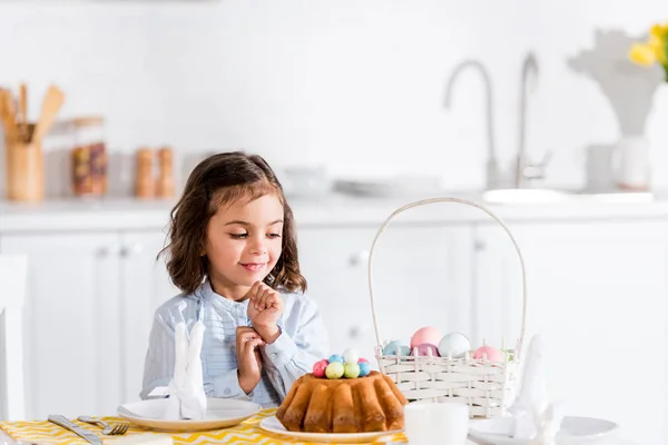 Симпатичный ребенок сидит за столом и смотрит на пасхальный торт и окрашенные яйца на кухне — стоковое фото