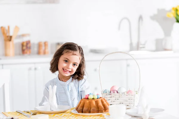 Niño adorable sentado a la mesa con pastel de Pascua y huevos pintados en la cocina - foto de stock