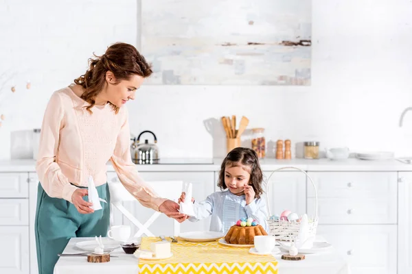 Mulher atraente dando filha dobrado guardanapo branco na cozinha — Fotografia de Stock