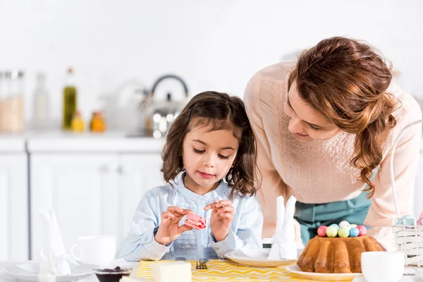 Mulher alegre olhando para a filha comendo macaroon na cozinha — Fotografia de Stock
