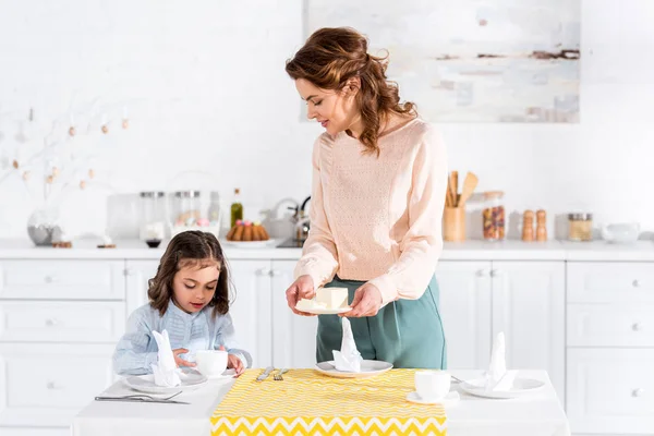 Mulher segurando pires com manteiga e olhando para a filha na cozinha — Fotografia de Stock