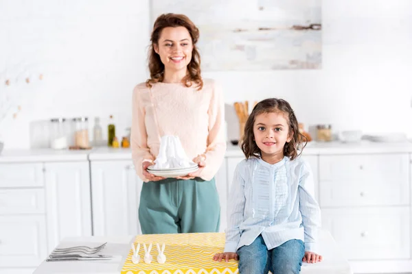 Woman with napkins looking at smiling little daughter in kitchen — Stock Photo