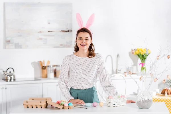 Lächelnde Frau mit Hasenohren und bemalten Ostereiern in der Küche — Stockfoto
