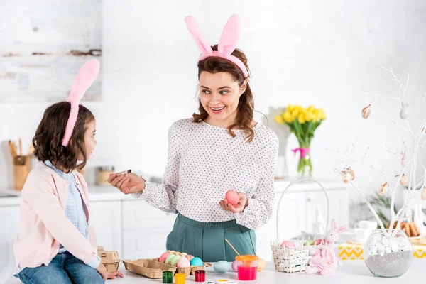 Mujer sonriente en orejas de conejo mirando a su hija mientras pinta huevo de Pascua en la cocina - foto de stock