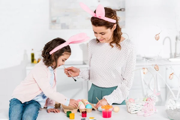 Madre e hija en orejas de conejo pintando huevos de Pascua en la cocina - foto de stock