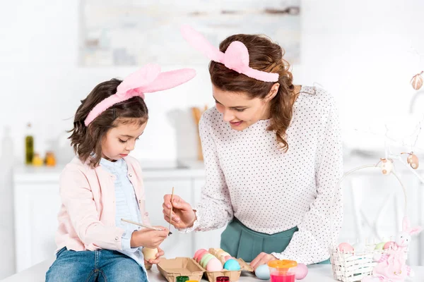Alegre madre e hija en orejas de conejo pintando huevos de Pascua en la cocina - foto de stock
