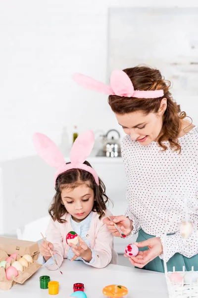Веселая мать и дочь в кроличьих ушах рисуют пасхальные яйца на кухне — стоковое фото