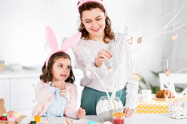 Smiling mother and daughter in bunny ears decorating easter tree in kitchen — Stock Photo
