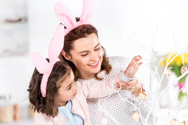 Joyful mother and daughter in bunny ears decorating easter tree together — Stock Photo