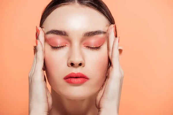 Beautiful trendy young woman with glitter makeup and eyes closed touching face isolated on coral — Stock Photo