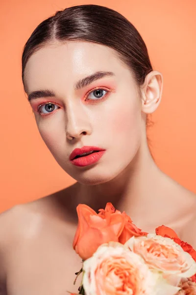 Hermosa chica con estilo mirando a la cámara y posando con flores de rosas aisladas en coral - foto de stock