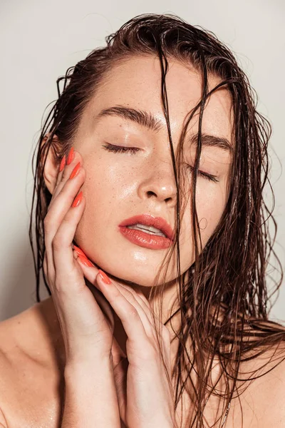 Bela jovem mulher com lábios de coral, olhos fechados e cabelos molhados tocando rosto no fundo cinza — Fotografia de Stock