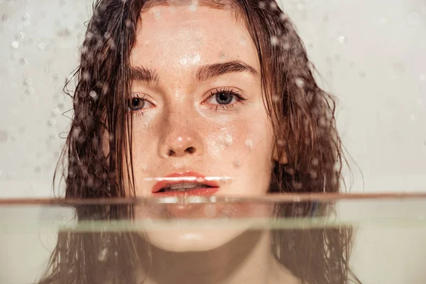 Schöne junge Frau mit Korallenlippen, die durch ein Glas mit Wassertropfen auf grau isoliert in die Kamera blickt — Stockfoto