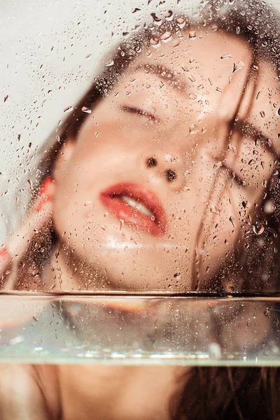 Beautiful young woman with coral lips posing through glass with water drops isolated on grey — Stock Photo