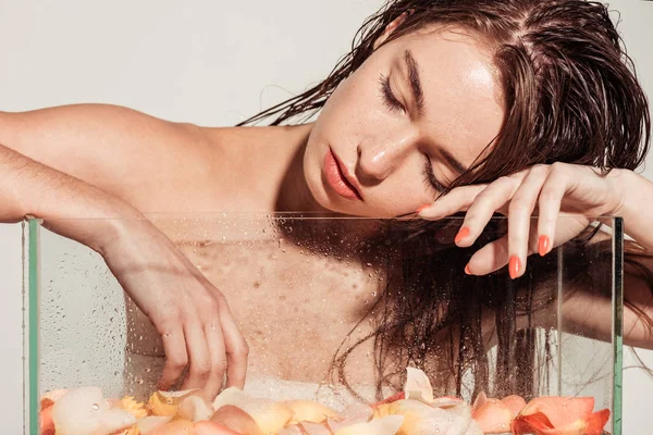 Beautiful young woman with coral lips and eyes closed posing near aquarium with rose petals isolated on grey — Stock Photo