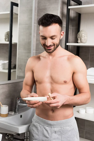 Веселий без сорочки чоловік дивиться на зубну пасту, тримаючи зубну щітку у ванній — стокове фото