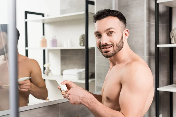 Hombre feliz sin camisa sosteniendo pasta de dientes y cepillo de dientes en el baño - foto de stock