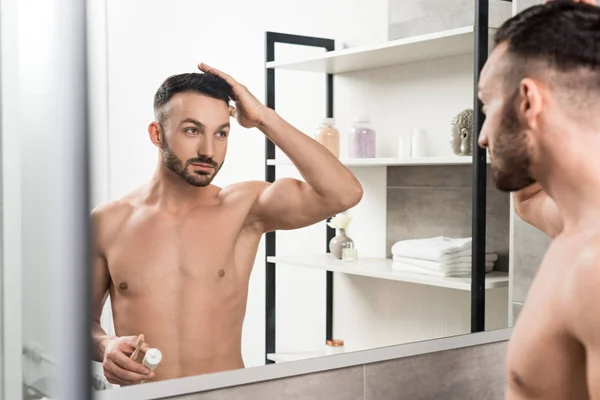 Schöner hemdloser Mann, der Haare berührt und Zahnbürste mit Zahnpasta in der Hand hält, während er im Badezimmer Spiegel anschaut — Stockfoto