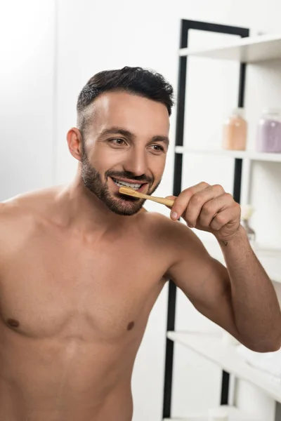Красивий без сорочки чоловік чистить зуби, дивлячись на дзеркало у ванній — стокове фото