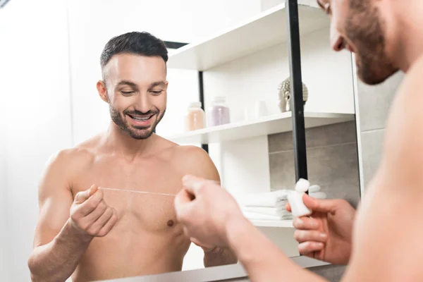 Весёлый бородатый мужчина смотрит на зубную нить, стоя возле зеркала в ванной комнате — стоковое фото