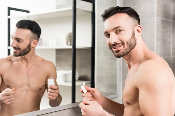 Весёлый бородатый мужчина держит зубную нить, стоя возле зеркала в ванной комнате — стоковое фото
