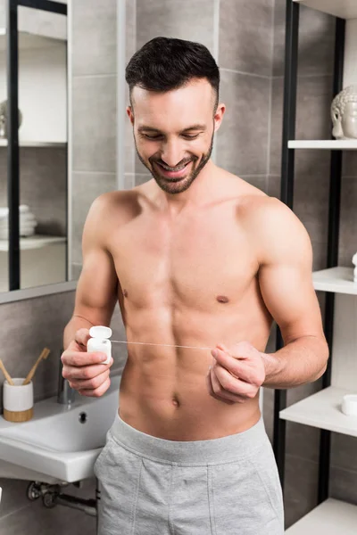 Hombre feliz sin camisa mirando el hilo dental mientras está de pie en el baño - foto de stock