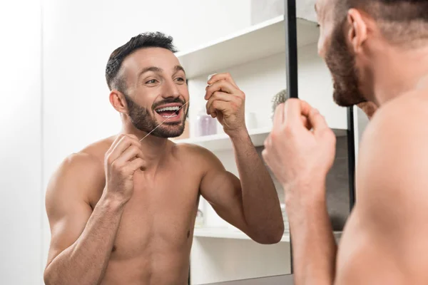 Счастливый мужчина без рубашки, смотрящий в зеркало, используя зубную нить в ванной комнате — стоковое фото