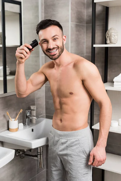 Alegre hombre sin camisa que sostiene trimmer mientras está de pie en el baño - foto de stock