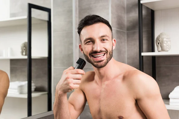 Веселый мужчина без рубашки с триммером рядом с лицом в ванной комнате — стоковое фото