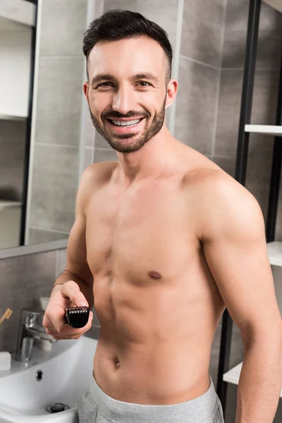 Hombre feliz sin camisa que sostiene trimmer mientras está de pie en el baño - foto de stock