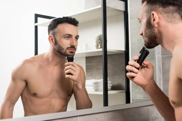 Handsome shirtless man holding trimmer while shaving face and looking in mirror in bathroom — Stock Photo