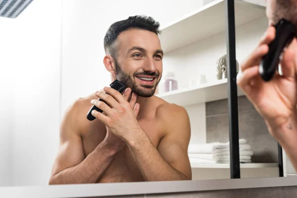 Homem sem camisa feliz segurando aparador ao raspar o rosto e olhando no espelho no banheiro — Fotografia de Stock
