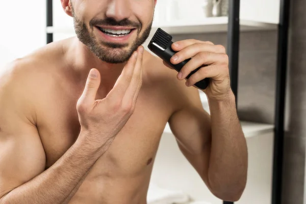 Cropped view of cheerful shirtless man holding trimmer while shaving face in bathroom — Stock Photo