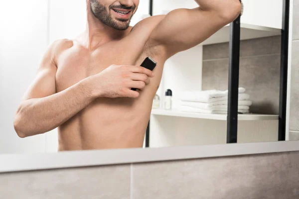 Vista recortada de hombre feliz sin camisa afeitándose la axila mientras está de pie cerca del espejo en el baño - foto de stock